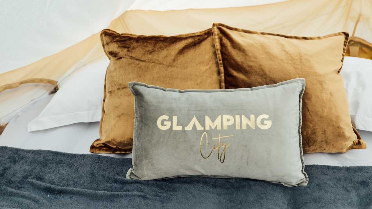 Drei Polster auf einem Bett. Die hinteren beiden Polster sind braun, das vordere Polster grau mit einer Aufschrift in Gold. "Glamping City" ist darauf abgedruckt, wobei das A in Glamping wie ein Zelt geformt ist.