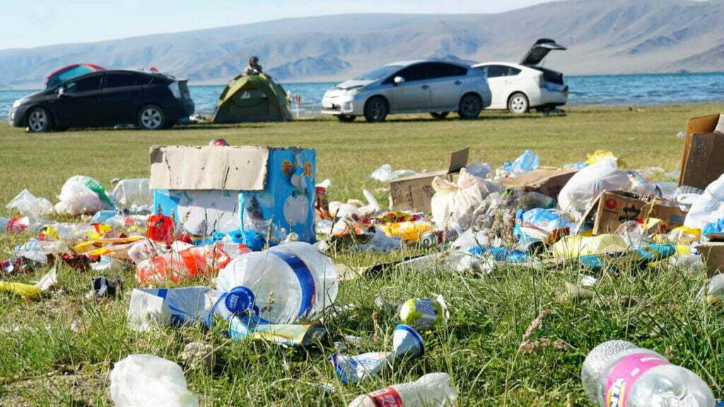 Ein Haufen Plastikmüll in einer Wiese. Im Hintergrund stehen Autos und ein Campingzelt direkt vor dem Wasser.