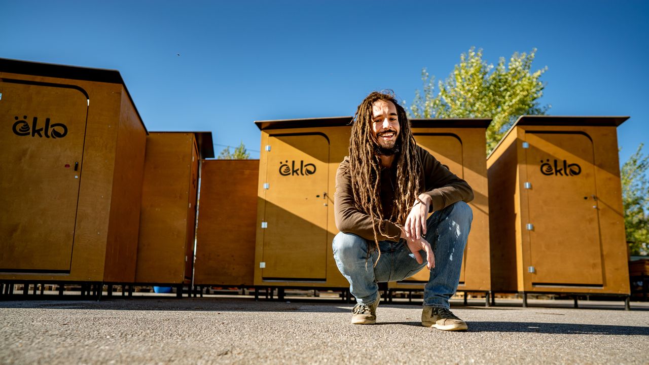 Niko, einer der Gründer von öKlo. Er hockt vor einigen öKlos auf dem Boden. Niko hat offene Dreadlocks, die bis zu seiner Hüfte reichen und lächelt in die Kamera.